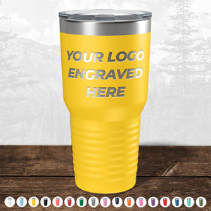 Yellow insulated tumbler from Kodiak Coolers with custom logo engraving space labeled "your logo engraved here" placed on a wooden surface with a blurred forest background.