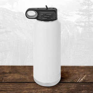 A white 32 oz Kodiak Coolers water bottle with a black flip top straw lid, laser engraved with the Wisconsin Green Bay Bulldogs logo, sits on wood with mountains in the background.