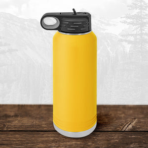 A Kodiak Coolers Wisconsin Luxemburg Casco Spartans 32 oz water bottle with a black flip top straw lid and laser-engraved design sits on a wooden surface, with a scenic mountain backdrop behind it.