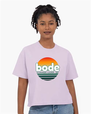 Woman wears the designerapp BODE Summer Camp 2026 "OG Logo" Women's Crop Top, featuring a sunset graphic and logo on soft, garment-dyed Comfort Colors 3023CL fabric in light purple.