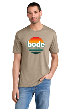 A man in a Printify 2026 Bode Summer Bootcamp DM130 Tri Blend Unisex T-Shirt, tan with "bode summer camp" and a sunset graphic on the front, stands with one hand in his pocket.