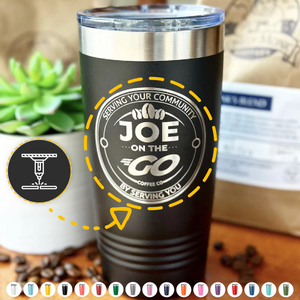 A Kodiak Coolers custom tumbler, laser engraved for added personalization, sits on a wooden surface surrounded by coffee beans, with a blurred background suggesting a cozy café setting.