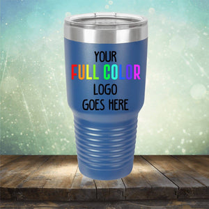 A blue Custom UV Printed Drinkware - ONE TIME SPECIAL OFFER with a stainless steel lid sits on a wooden surface. The text "YOUR FULL COLOR LOGO GOES HERE" is printed on the tumbler, resembling the polished look of coffee mugs. The background features a bokeh effect with green and yellow hues. This exclusive product is brought to you by Kodiak Coolers.