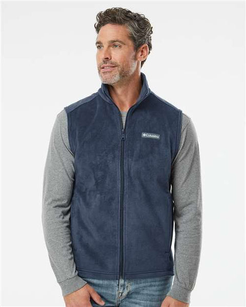 A man donning a Columbia Steens Mountain Fleece Vest in navy, crafted from polyester fleece, and a gray long-sleeve shirt, stands against a plain white background. Ideal for outdoor activities.