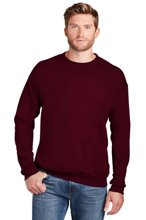 A man wearing a Hanes Ultimate 100% Cotton Crewneck Sweatshirt F260 in maroon and blue jeans stands with one hand in his pocket against a white background.