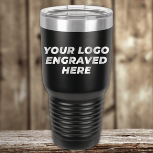 A black Kodiak Coolers tumbler with a clear lid sits on a wooden surface, featuring "YOUR CUSTOM COMPANY LOGO"—a perfect Realtor closing gift for clients or an addition to your personalized drinkware collection.