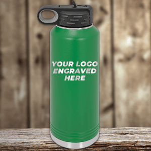 A green metal water bottle by Kodiak Coolers, with a black lid on a wooden surface, features "YOUR LOGO ENGRAVED HERE" in white—ideal as Realtor closing gifts or promotional drinkware with your custom company logo.